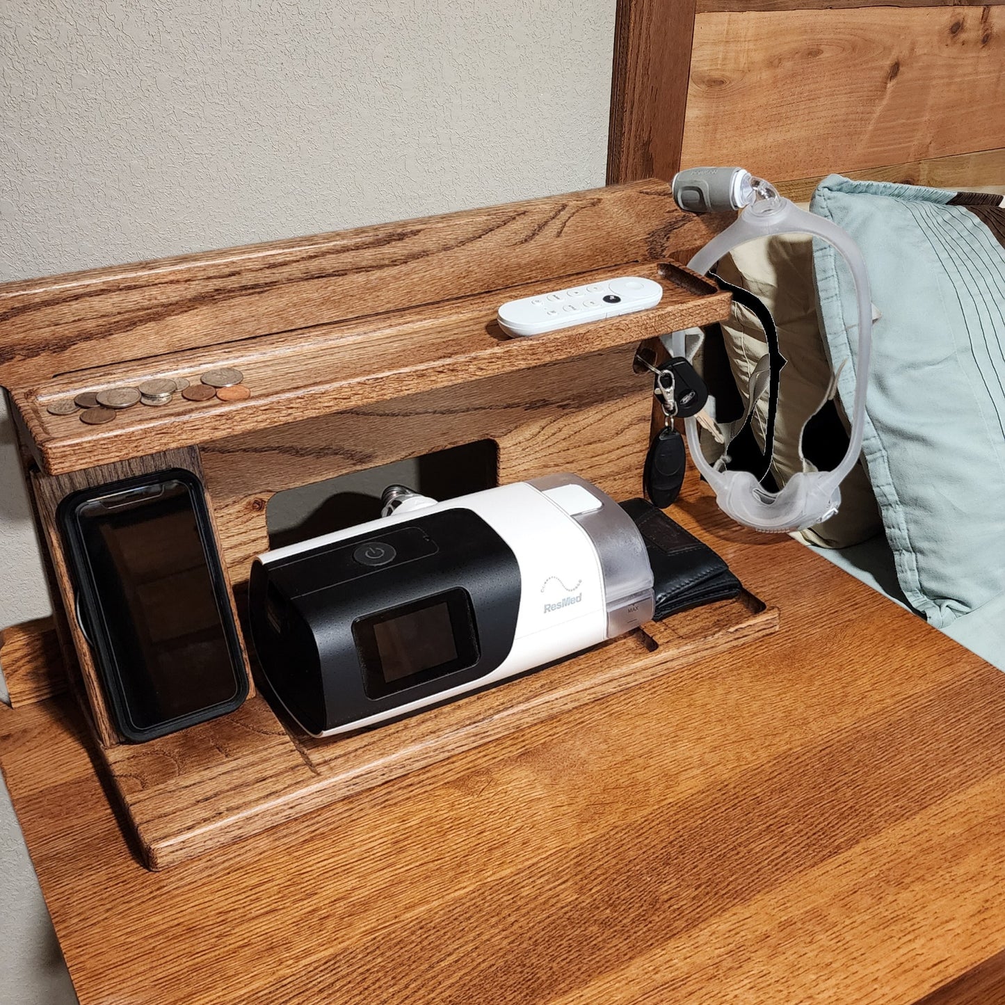 A CPAP Smart Stand that is displayed next to a bed holding the CPAP machine, wirelessly charging the phone and watch and has a few items in the catch-all trays. The mask is also hanging from the stand to keep it off the nightstand and floor. Items on the stand are very organized.