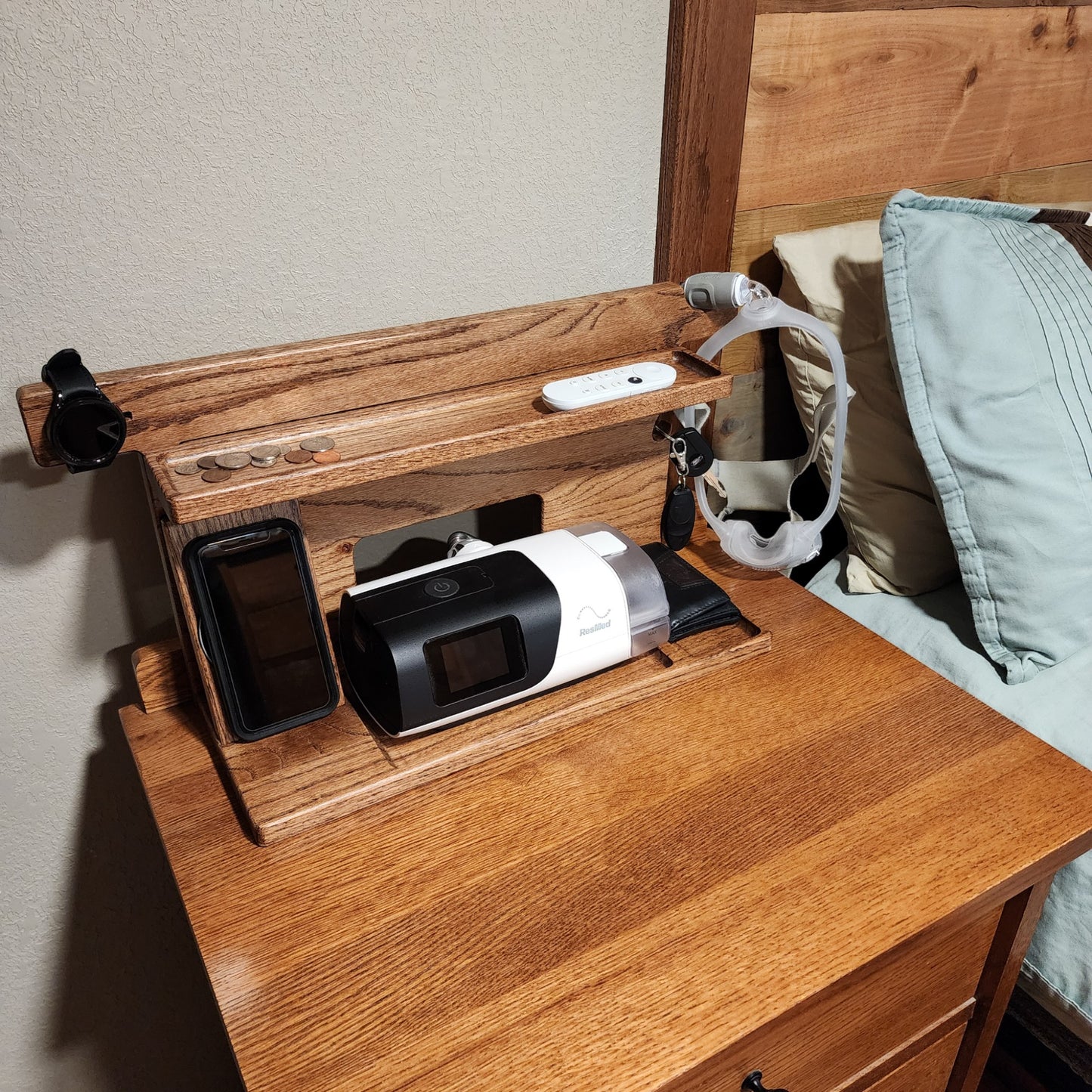 A CPAP Smart Stand that is displayed next to a bed holding the CPAP machine, wirelessly charging the phone and watch and has a few items in the catch-all trays. The mask is also hanging from the stand to keep it off the nightstand and floor. Items on the stand are very organized.