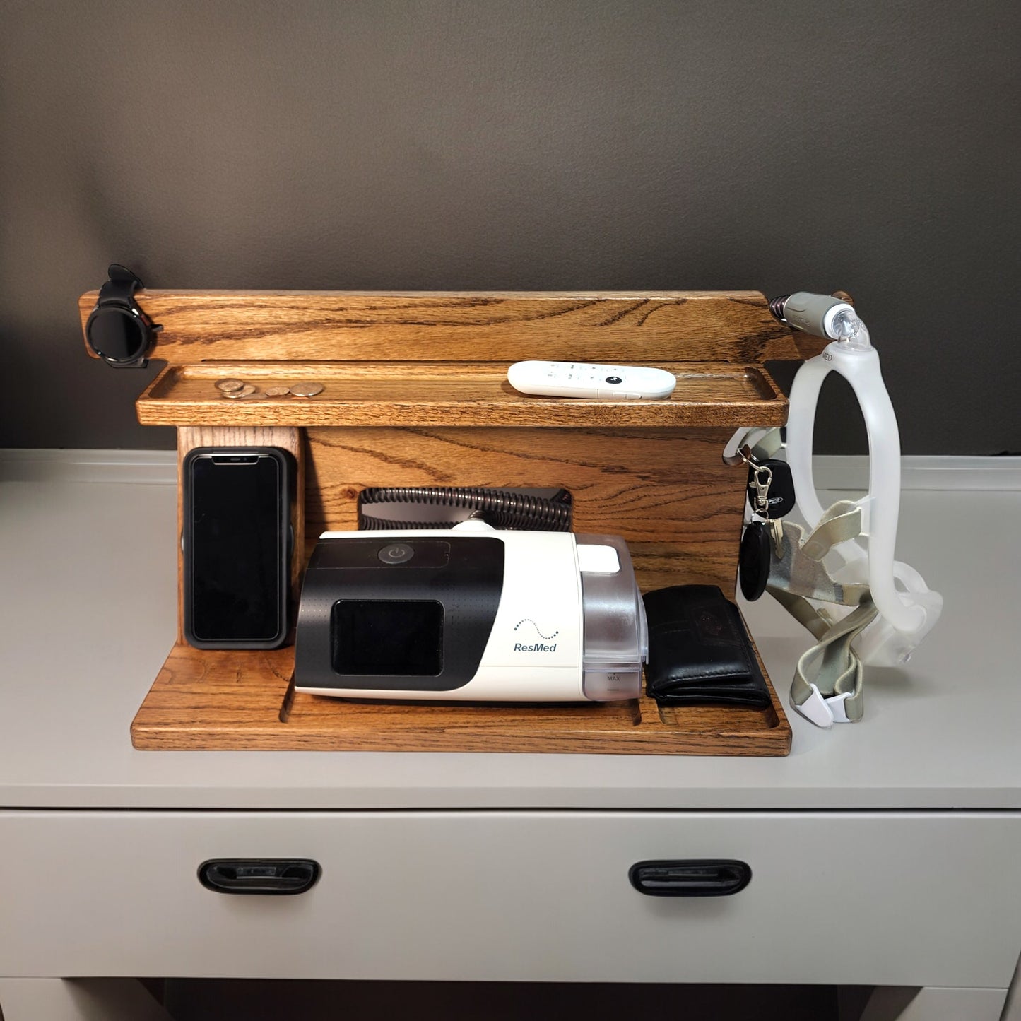A CPAP Smart Stand that is charging a smart watch and charging a smart phone. The stand is holding the CPAP machine, change, TV remote control, car keys and a wallet. The mask is hanging from the stand to keep it off the nightstand and floor. The stand has three catch-all trays and is made from solid oak wood.