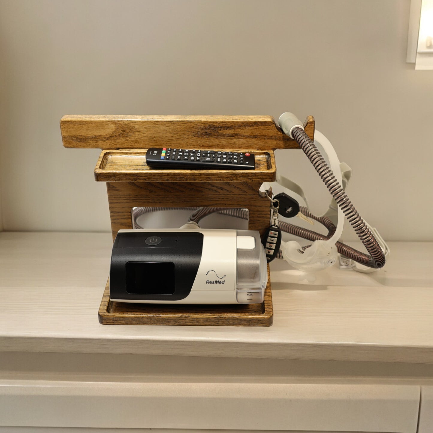 A CPAP Stand that has the CPAP inserted and mask hanging from the stand. The stand is made by Custom Cave works and is made from solid oak wood. The stand is also holding car keys and a TV remote.