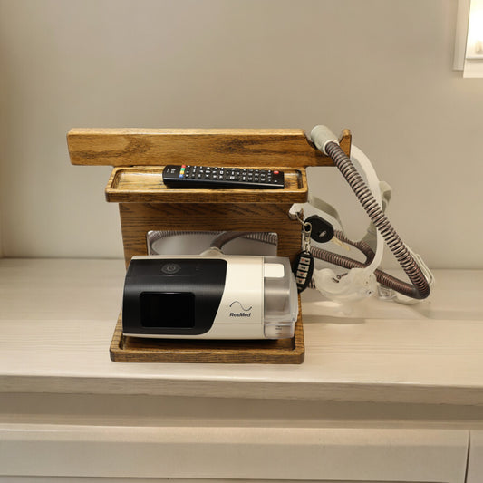 A CPAP Stand that has the CPAP inserted and mask hanging from the stand. The stand is made by Custom Cave works and is made from solid oak wood.  The stand is also holding car keys and a TV remote.