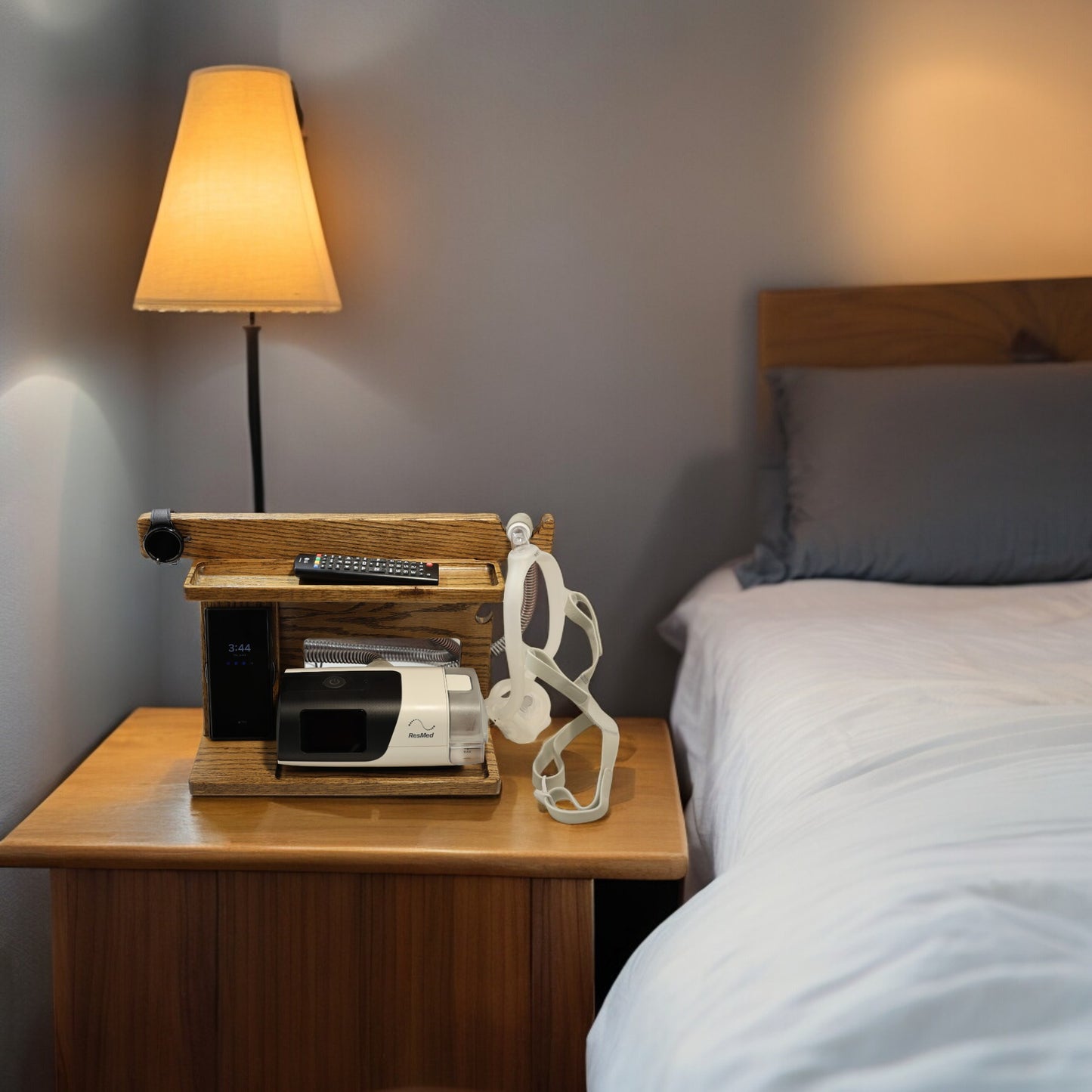 A CPAP Smart Stand that is charging a smart watch and charging a smart phone. The stand is holding the CPAP machine, TV remote control, and has room for other items. The mask is hanging from the stand to keep it off the nightstand and floor. The stand has two catch-all trays and is made from solid oak wood.