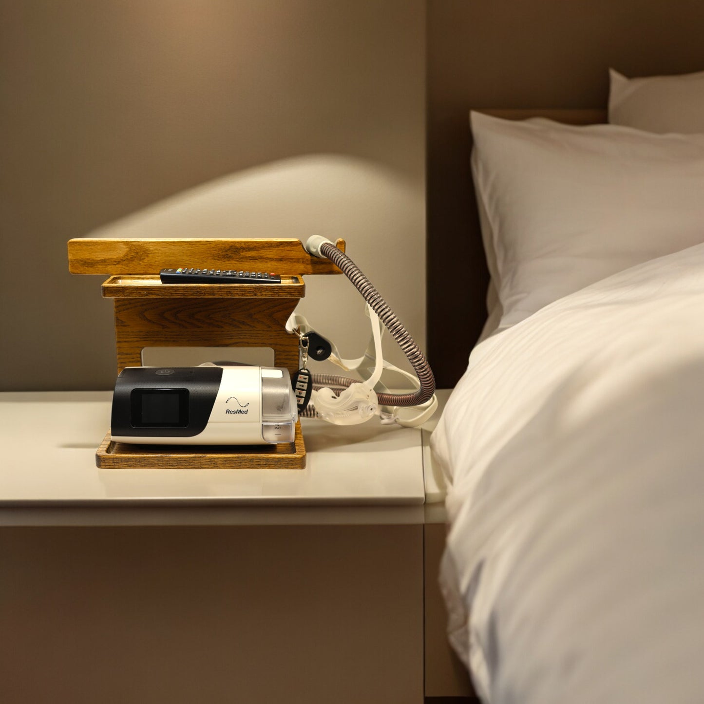 A CPAP Stand next to the comfortable bed, The stand is holding the CPAP machine, TV remote control, and has room for other items. The mask is hanging from the stand to keep it off the nightstand and floor. The stand has two catch-all trays and is made from solid oak wood.